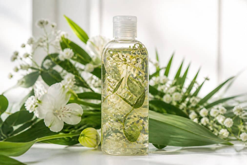 Primo piano di una bottiglia di shampoo trasparente con ingredienti naturali visibili al suo interno, circondata da foglie verdi fresche e fiori bianchi su sfondo bianco minimalista