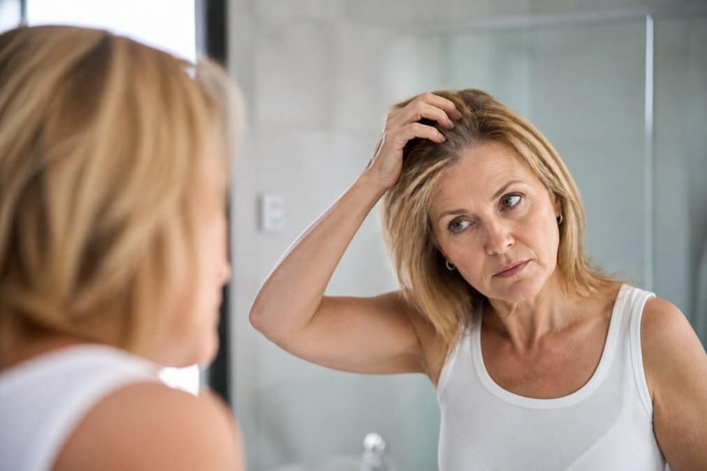 Donna di mezza età che esamina i propri capelli fini allo specchio in bagno luminoso, espressione consapevole e riflessiva