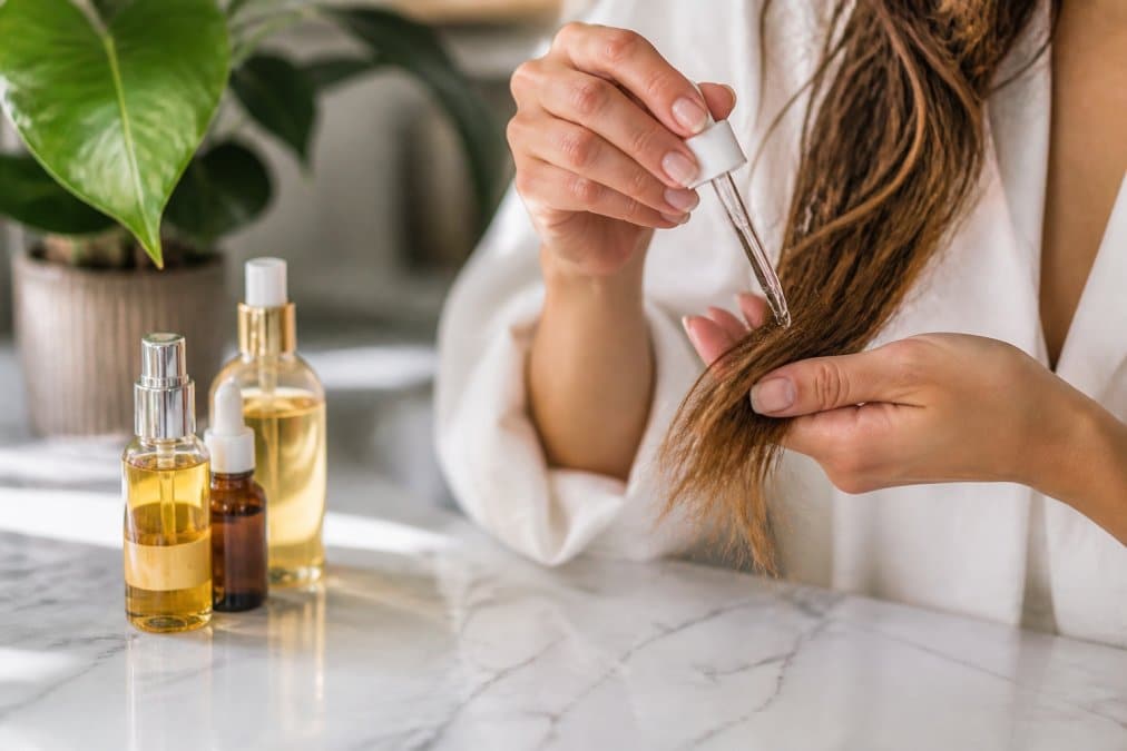 Mani che applicano un siero o trattamento liquido sulle punte dei capelli, con bottiglie di prodotto riparatore in primo piano su superficie di marmo