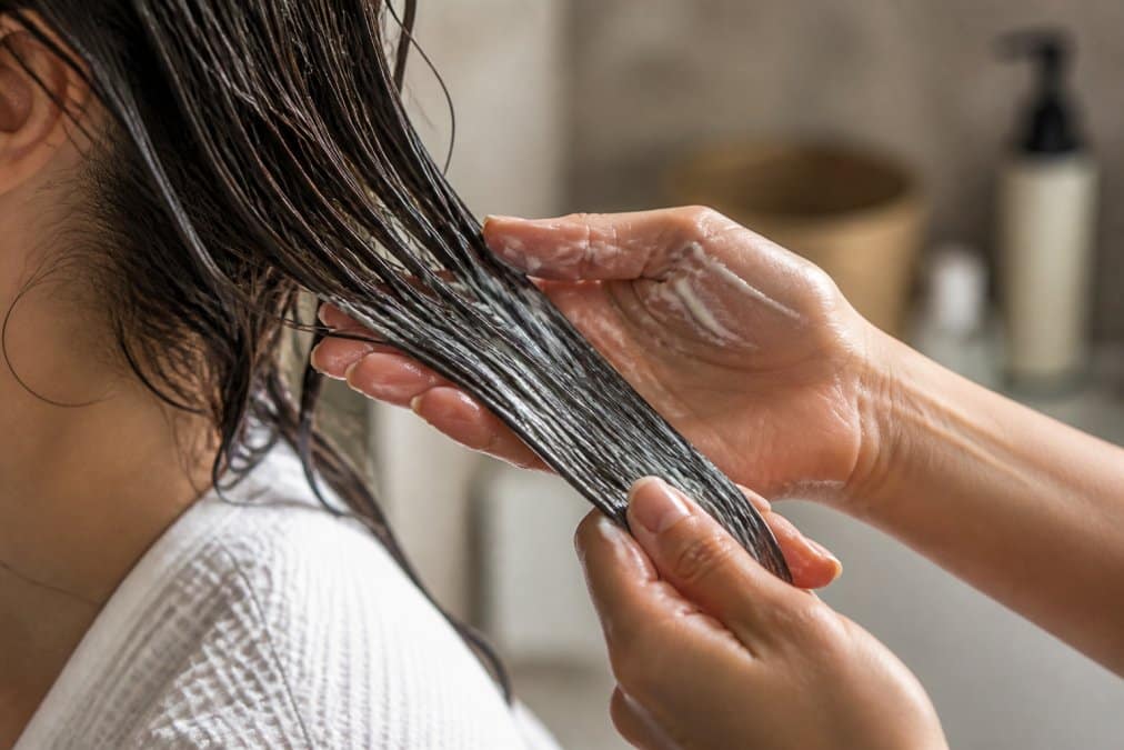 Close-up delle mani che applicano la maschera bio su una ciocca di capelli, mostrando la distribuzione uniforme dalla radice alle punte