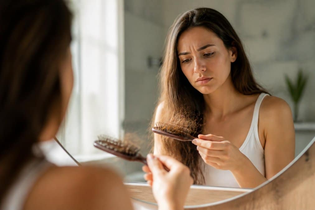 Donna che osserva i capelli caduti nel pettine con luce naturale che illumina i capelli e il cuoio capelluto, seduta davanti a uno specchio in bagno luminoso
