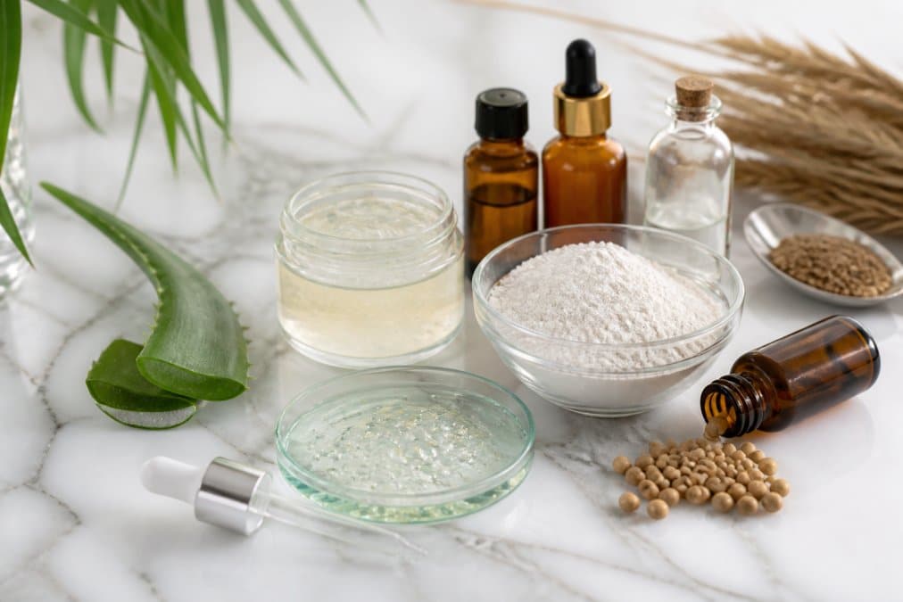 Bottles and containers of anti-aging products with visible natural ingredients, hyaluronic acid and keratin clearly displayed on a white marble surface